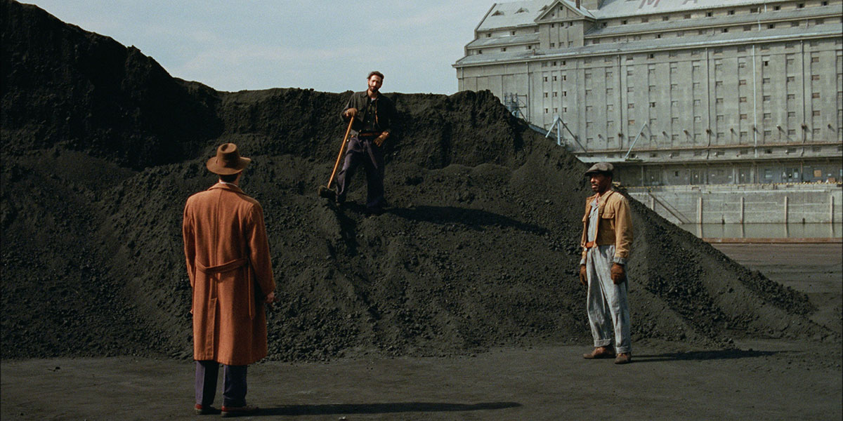 A still from The Brutalist with three men in old time-y attire standing in a triangle formation near a dirt hill